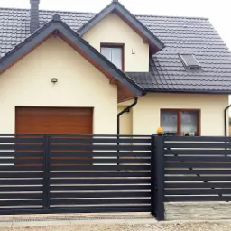 Nowoczesna, pozioma, czarna brama metalowa z przesuwnym skrzydłem na tle domu z brązową bramą garażową i beżową elewacją.