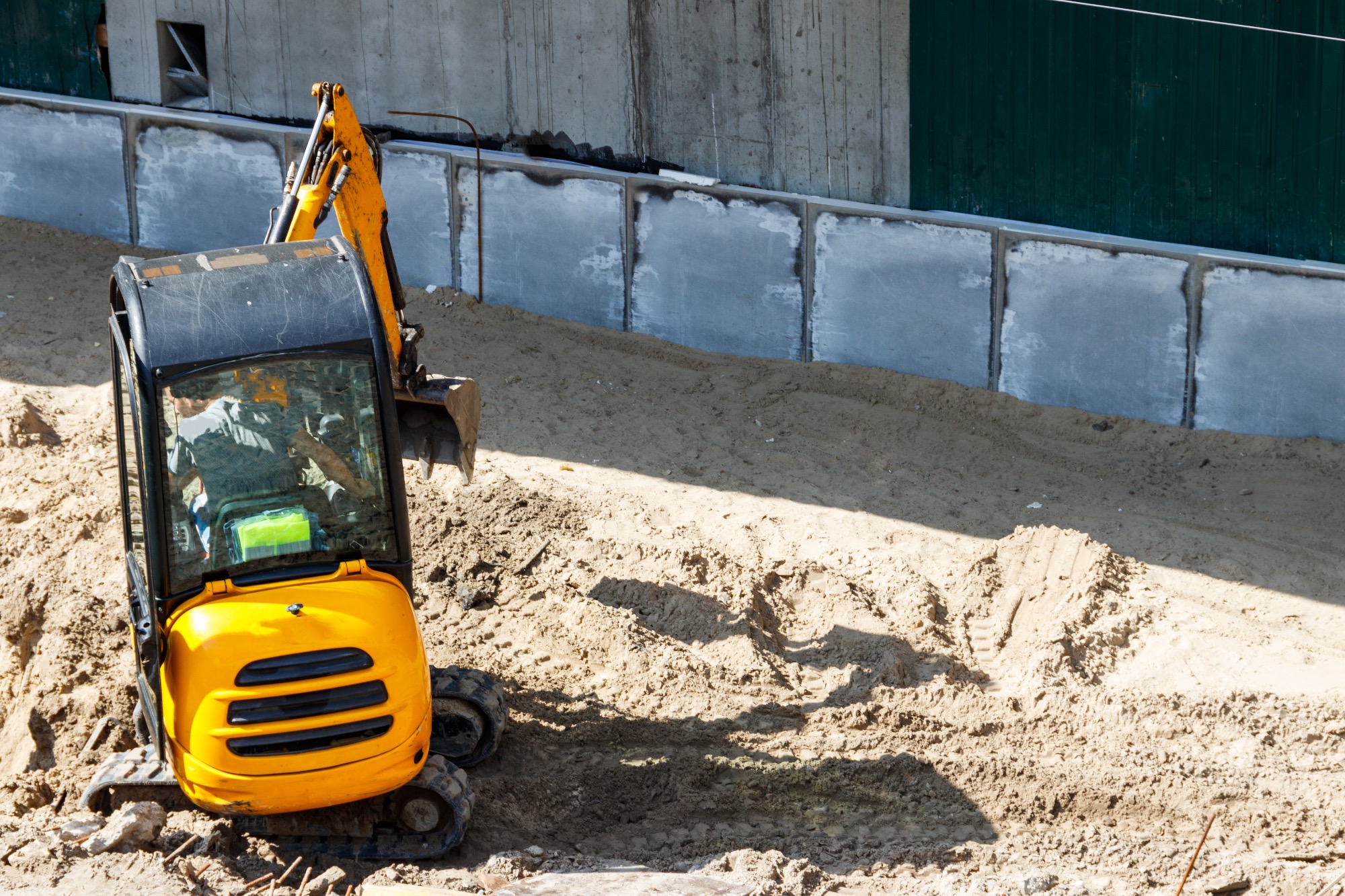 Żółta mini koparka na placu budowy, widoczny operator w kabinie, w tle betonowe ściany i wykopany teren