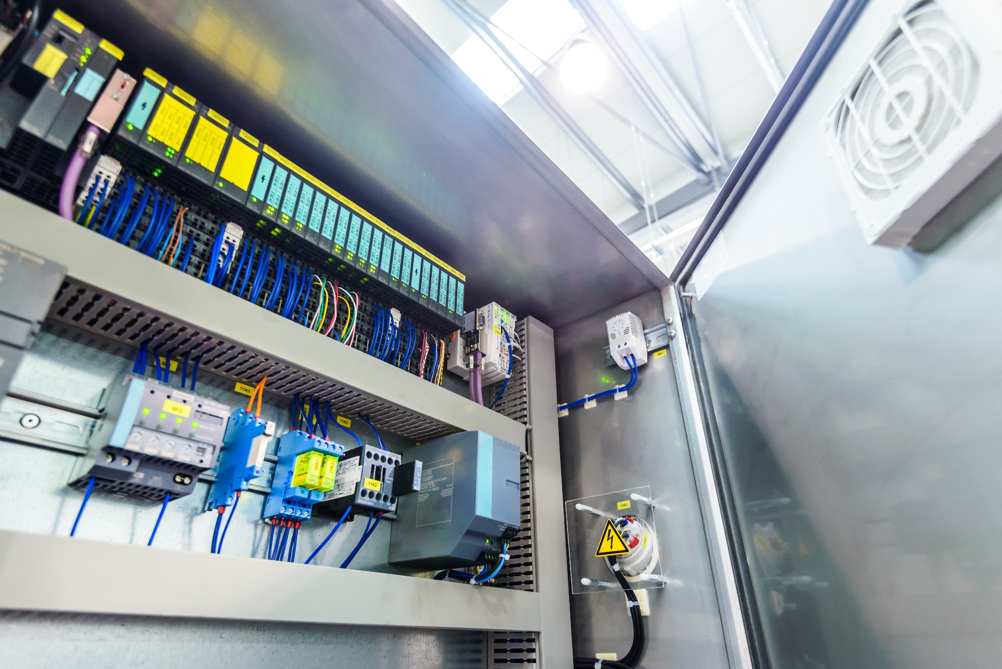 Otwarta szafa sterownicza z modułami PLC, bezpiecznikami i okablowaniem, widoczny wentylator chłodzący i ostrzeżenie o napięciu.