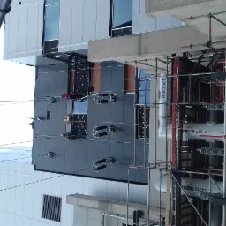 Pracownicy na wysokościach, zabezpieczeni linami, montują elementy na elewacji budynku. Widoczny dźwig i rusztowania. Szara elewacja z czarnymi akcentami.