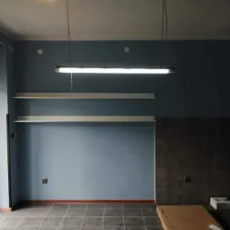 Pomieszczenie po malowaniu ścian na kolor niebieski, z zamontowanymi półkami i oświetleniem jarzeniowym, płytki na podłodze i dolnej części ściany.