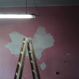Fach-Nid - Różowa ściana w trakcie przygotowania do malowania, z widocznymi obszarami zagruntowanymi i drewnianą drabiną opartą o ścianę, oświetlona jarzeniówką