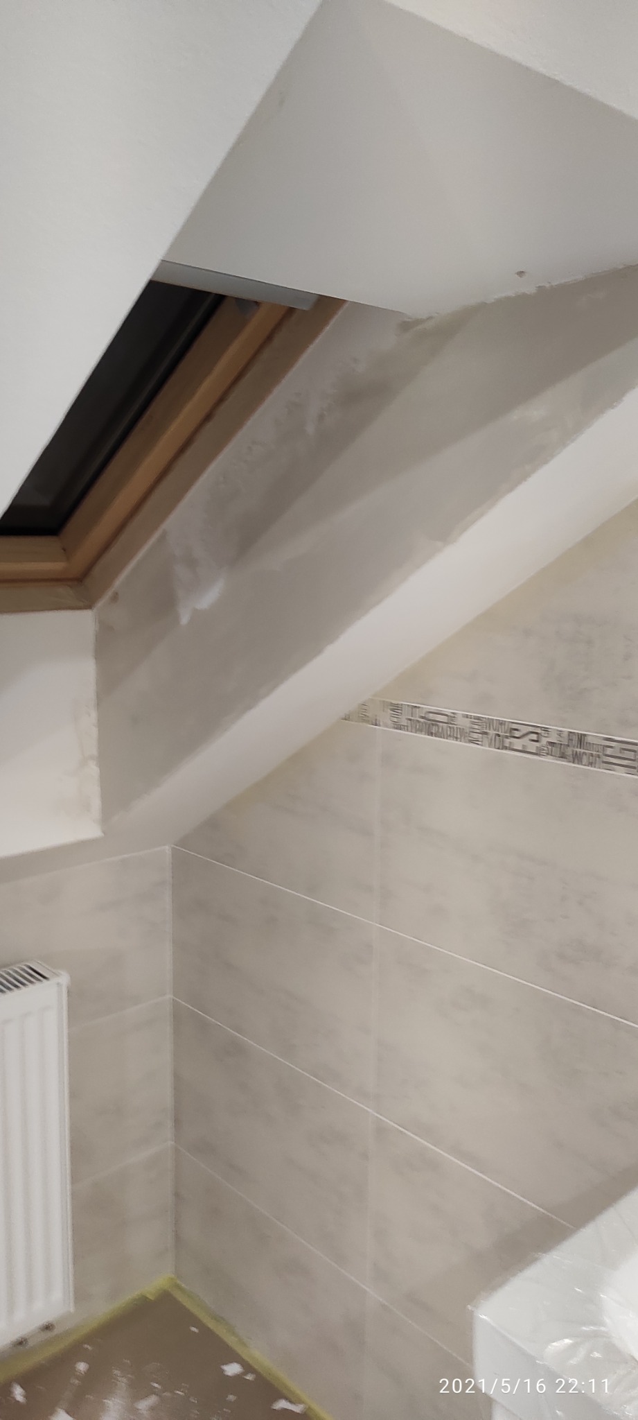 Poddasze z oknem dachowym w trakcie prac wykończeniowych, widoczne płytki na ścianie, częściowo zaszpachlowana ściana i zabezpieczona podłoga.