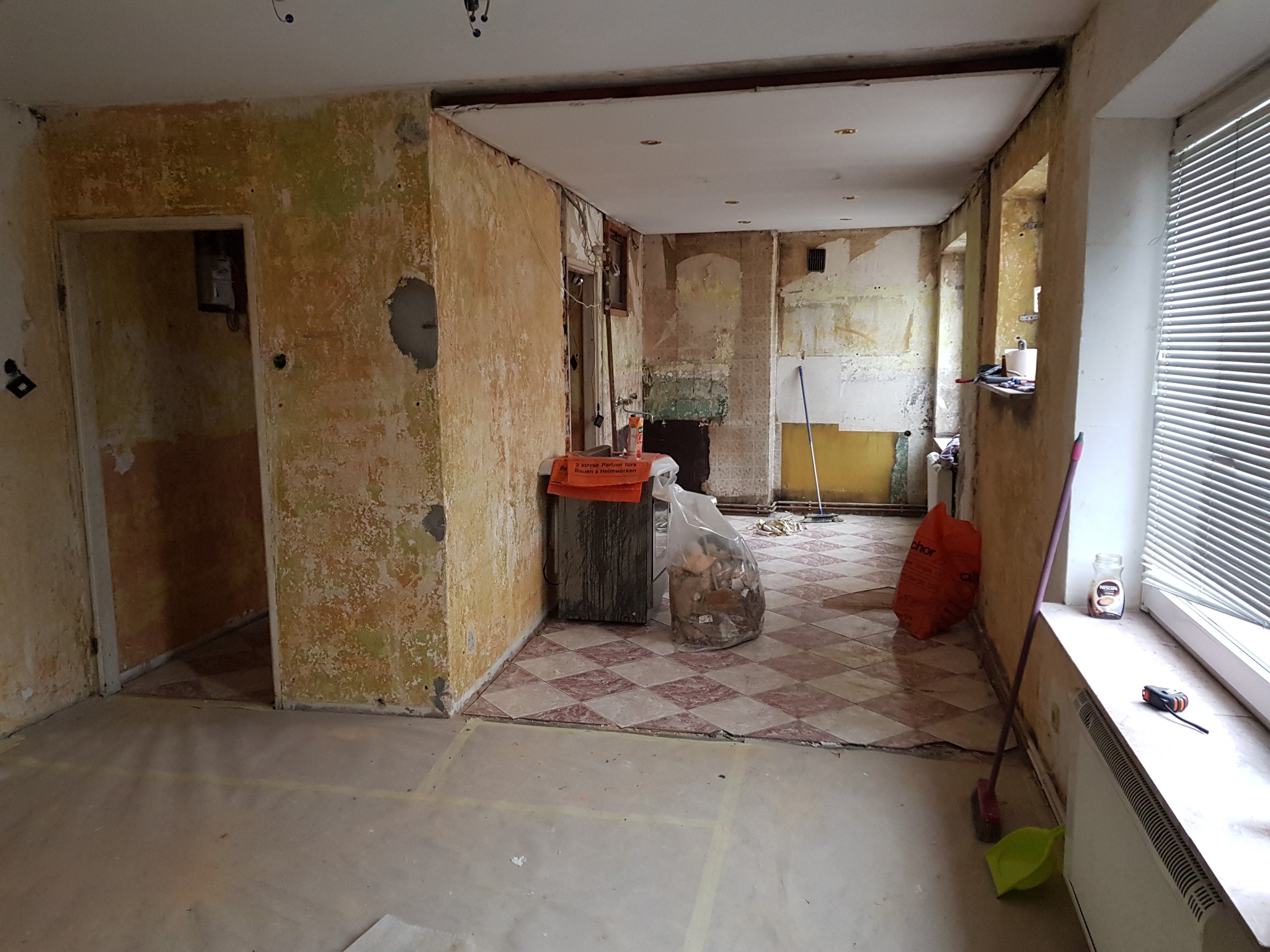 Wnętrze pokoju w trakcie prac przygotowawczych do remontu, ściany z resztkami starej farby, podłoga częściowo zabezpieczona folią, widoczne narzędzia i worki z odpadami.