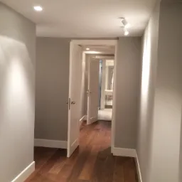 Korytarz z drewnianą podłogą, szarymi ścianami i białymi listwami przypodłogowymi, z widokiem na łazienkę przez otwarte drzwi, oświetlony halogenami.