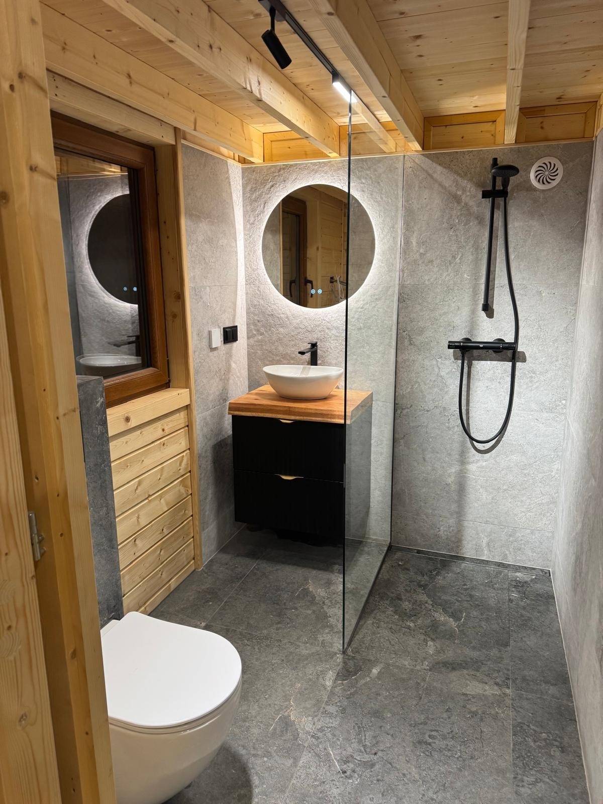 Nowoczesna łazienka w drewnie i szarości. Umywalka nablatowa, okrągłe lustro z podświetleniem LED, kabina prysznicowa typu walk-in, drewniane wykończenie ścian.