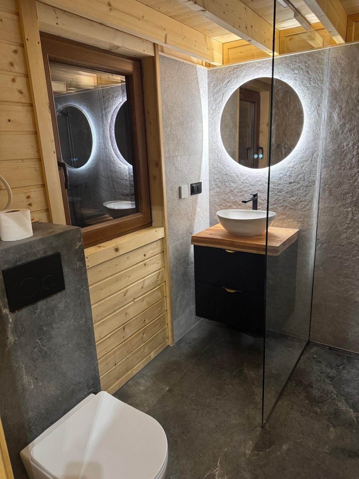 Drewniana łazienka z szarą armaturą: okrągłe lustro z podświetleniem, umywalka na szafce, toaleta. Widoczne drewniane panele i szare płytki imitujące kamień.