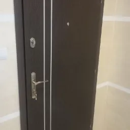 Ciemnobrązowe drzwi wejściowe z dwoma pionowymi, białymi paskami i srebrną klamką, zamontowane w jasnym przedpokoju z beżowymi ścianami.