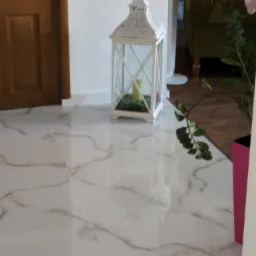 Błyszcząca, biała podłoga z marmurkowym wzorem, dekoracyjna latarenka ze świecą w środku, fragment zielonej sofy i rośliny doniczkowej w różowej osłonce w tle.