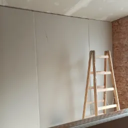 Montaż płyt kartonowo-gipsowych na ścianie, widoczna drewniana drabina i fragment ściany z cegły