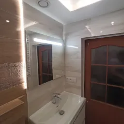 Nowoczesna łazienka z umywalką w szafce, lustrem z podświetleniem LED i płytkami imitującymi drewno na ścianie, widoczny fragment drzwi z szybą.