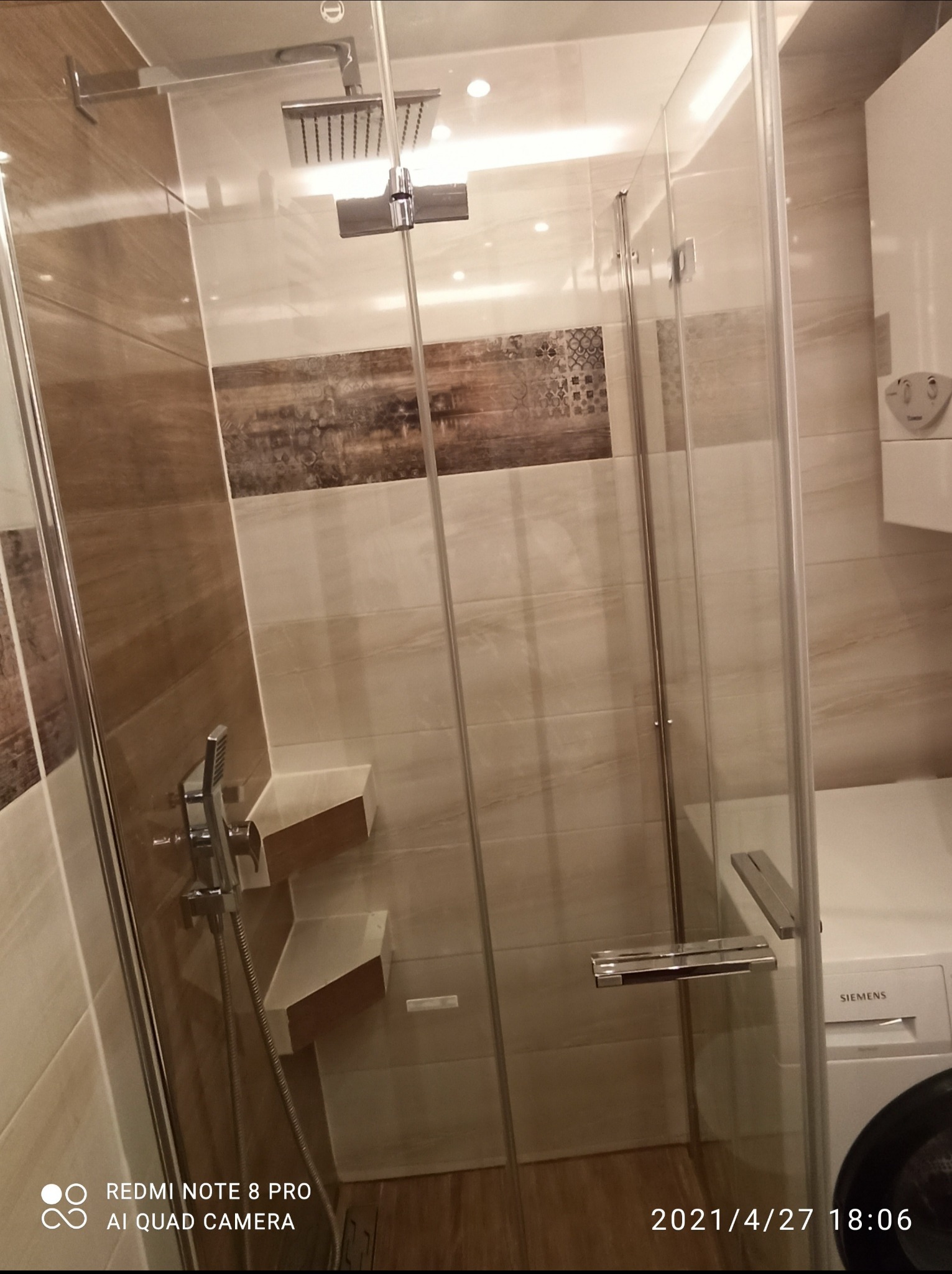 Nowoczesna kabina prysznicowa z deszczownicą i półkami wewnątrz, wykończona płytkami w odcieniach beżu i brązu, z widocznym bojlerem i pralką Siemens w tle.