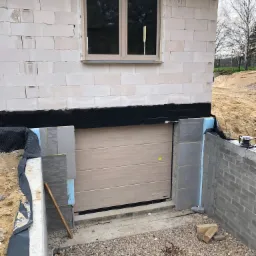 Montaż jasnej bramy garażowej segmentowej w surowym stanie budynku, widoczne bloczki betonowe, okno i izolacja termiczna. Ukończona wnęka z drobnym żwirem.