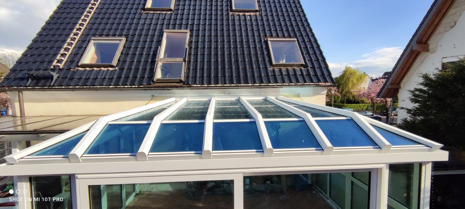 Zadaszenie tarasu z białymi profilami i szklanym dachem, widok z góry na tle dachu domu z oknami połaciowymi i kwitnących drzew.