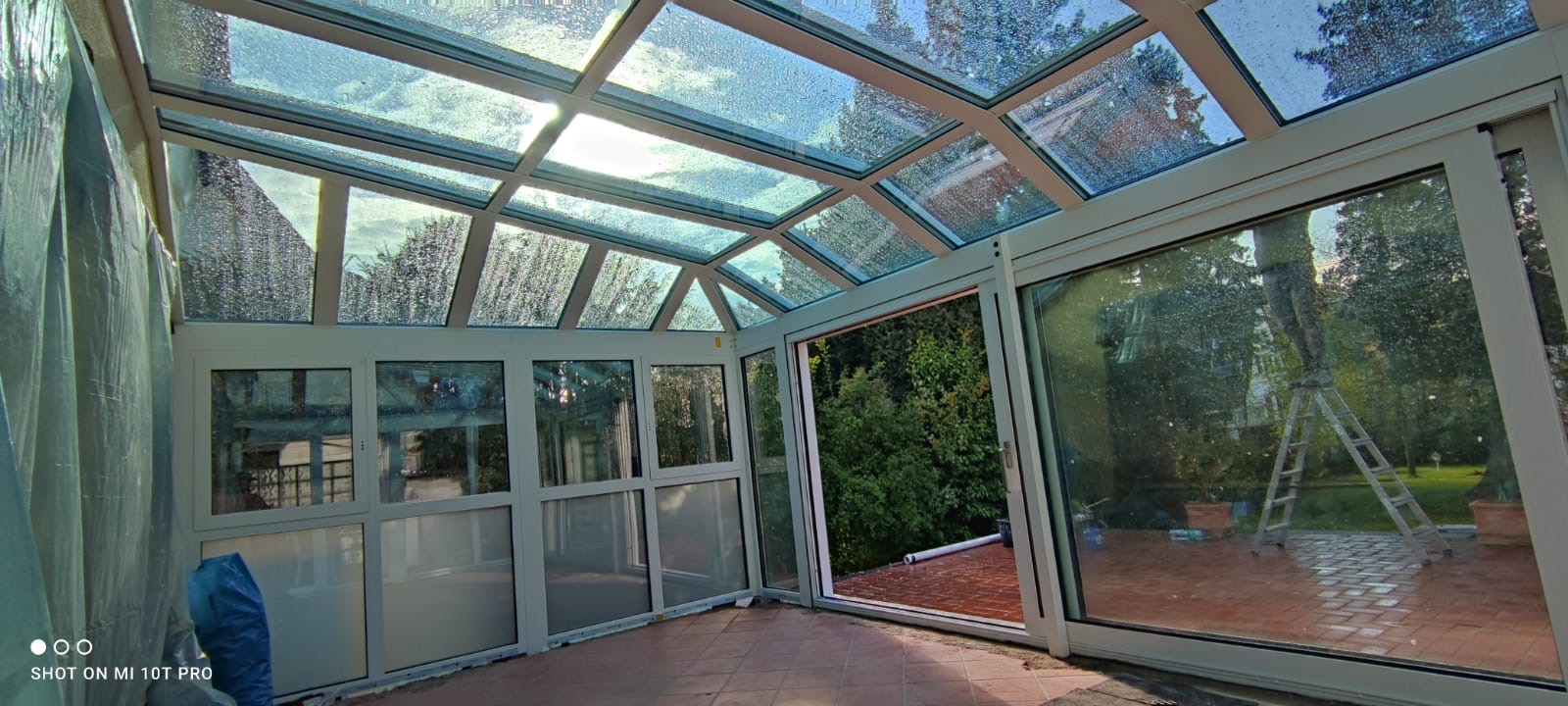 Szklana weranda z oknami aluminiowymi, widoczne krople deszczu na szybach oraz drabina na zewnątrz.