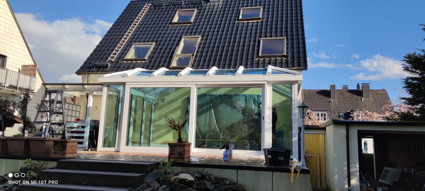 Nowoczesny ogród zimowy z białymi ramami, przeszklonym dachem i przesuwnymi drzwiami, widok z zewnątrz na tle domu z dachówką i oknami dachowymi, wewnątrz widoczne narzędzia i osłona.