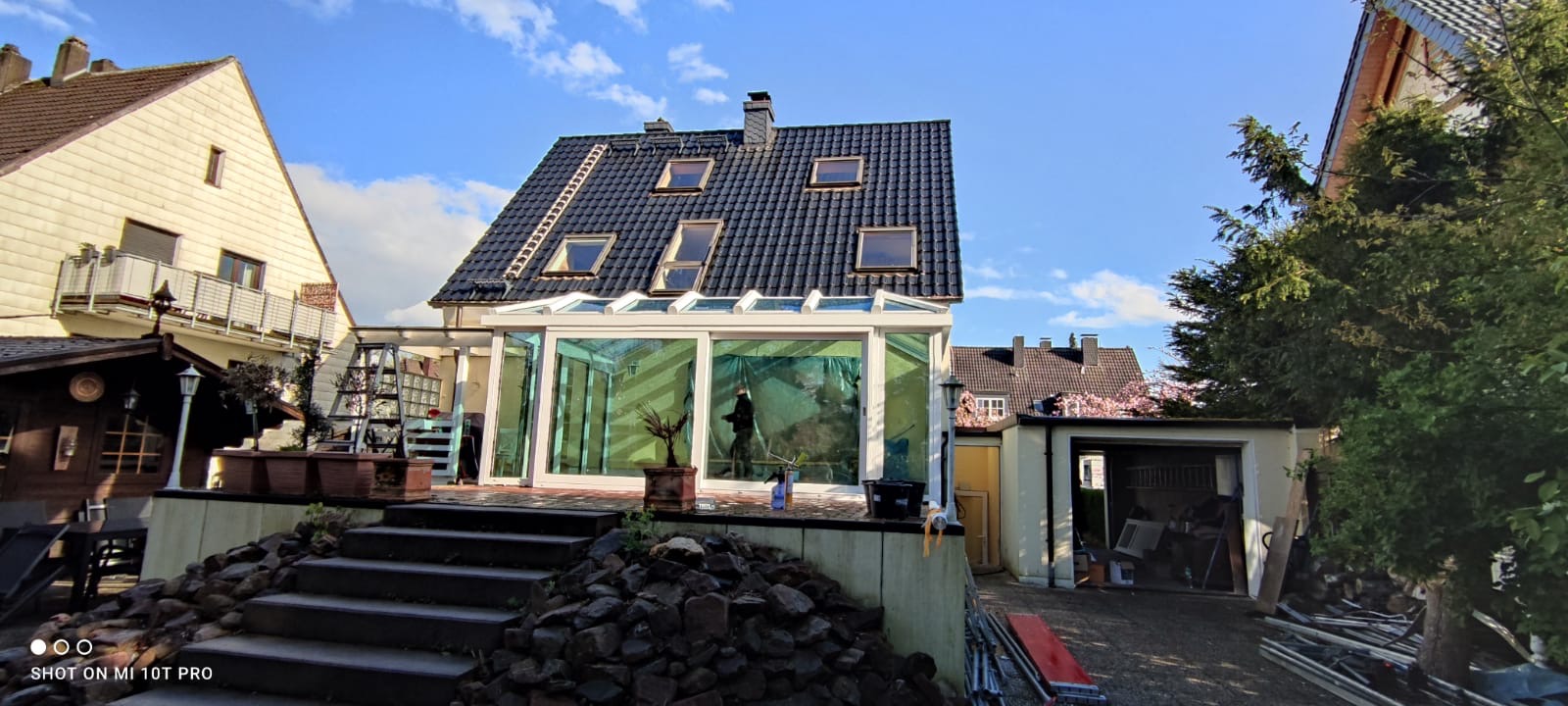 Widok z dołu na przeszkloną oranżerię z białymi ramami, dobudowaną do domu z ciemną dachówką i oknami dachowymi, na dachu drabina, wewnątrz oranżerii widoczna osoba, na pierwszym planie kamienne...