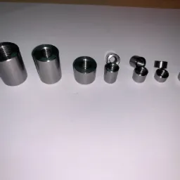 Różnej wielkości, precyzyjnie wykonane, metalowe tuleje gwintowane ułożone w rzędzie na białym tle, ukazujące wewnętrzne gwinty. Największa tuleja ma kształt lekko stożkowy.