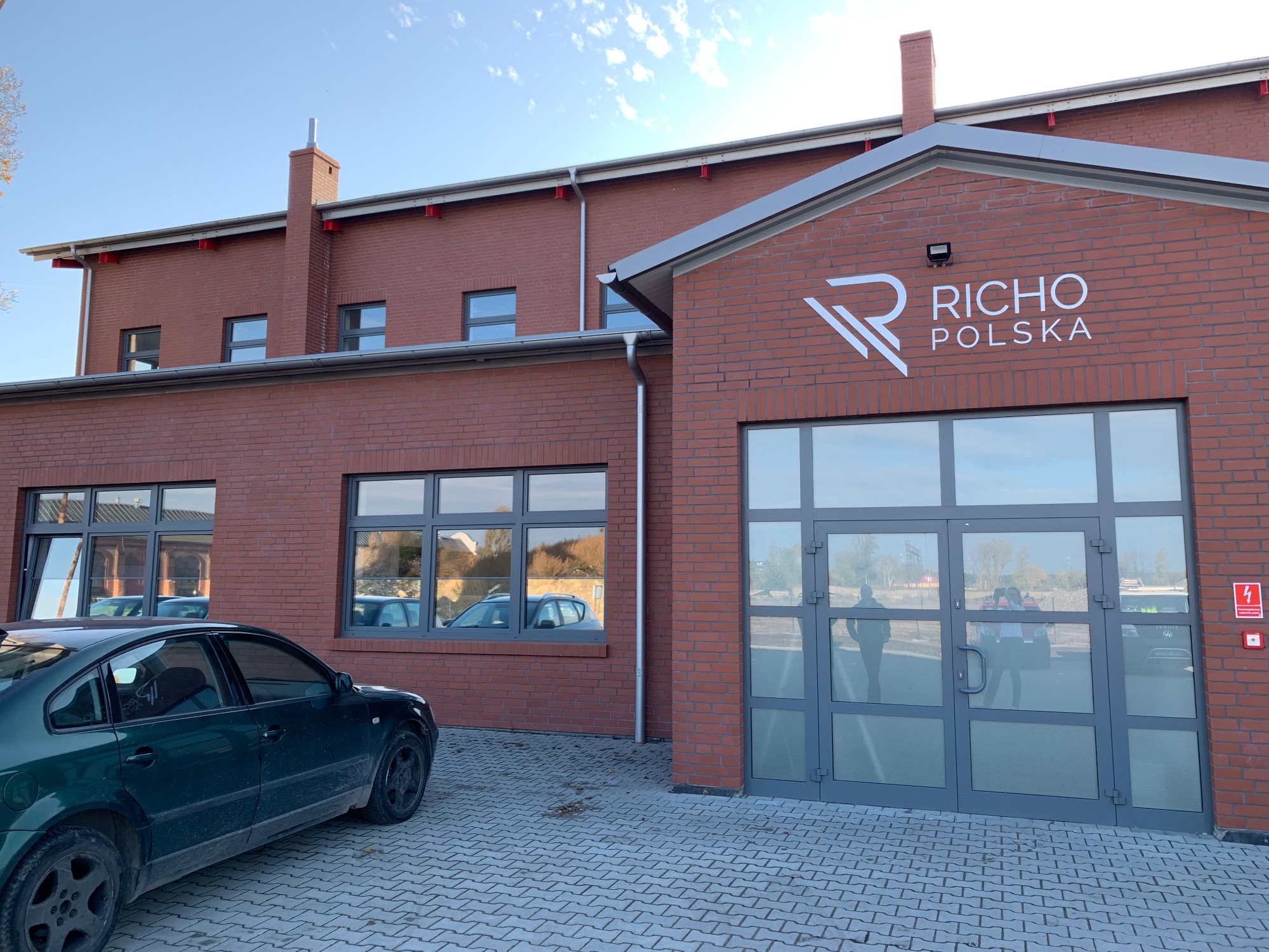 Budynek firmy z czerwonej cegły z logo 'RICHO POLSKA' nad szklanymi drzwiami wejściowymi, na pierwszym planie zaparkowany ciemnozielony samochód.