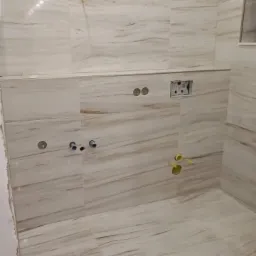 Ściany i podłoga wyłożone płytkami imitującymi marmur w odcieniach bieli i beżu, widoczne otwory na instalacje sanitarne, etap prac wykończeniowych.