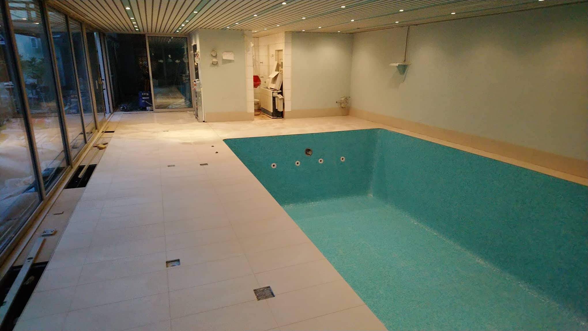 Wnętrze basenu w trakcie wykańczania: turkusowa niecka, jasne płytki na podłodze z miejscami na odpływy, duże okna przesuwne z widokiem na zewnątrz, drewniany sufit z oświetleniem punktowym.