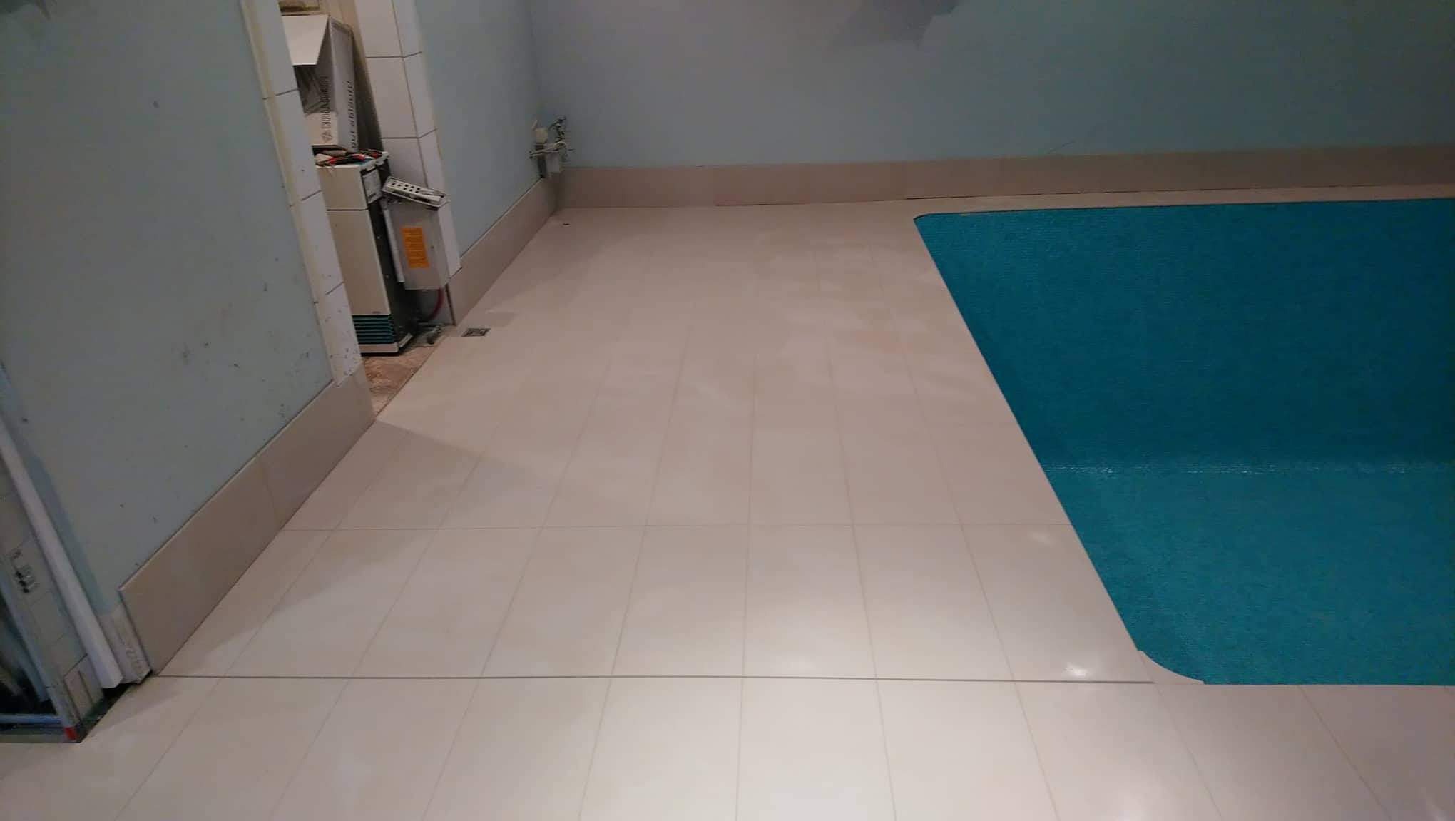 Wyłożona jasnymi płytkami podłoga łazienki z fragmentem zabezpieczonej folią wanny lub brodzika, widoczne fragmenty instalacji elektrycznej i hydraulicznej.