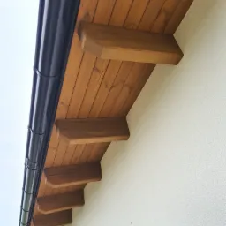 Drewniana podbitka dachowa z widocznymi krokwiami i czarną rynną, przymocowana do jasnej elewacji budynku.