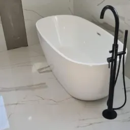 Wolnostojąca wanna w nowoczesnej łazience z białymi marmurowymi płytkami i czarną armaturą.