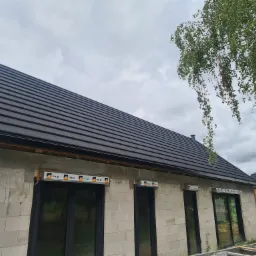 Niedokończony dom murowany z czarnym dachem z dachówki ceramicznej, ściany z białego betonu komórkowego, okna z roletami zewnętrznymi w trakcie montażu, widoczne rusztowanie i drzewo brzozy na tle...