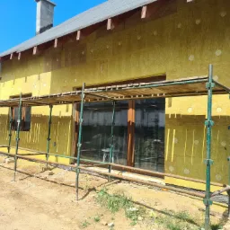 Dom w trakcie ocieplania elewacji wełną mineralną, widoczne rusztowanie, fragment dachu i komina, słoneczny dzień.