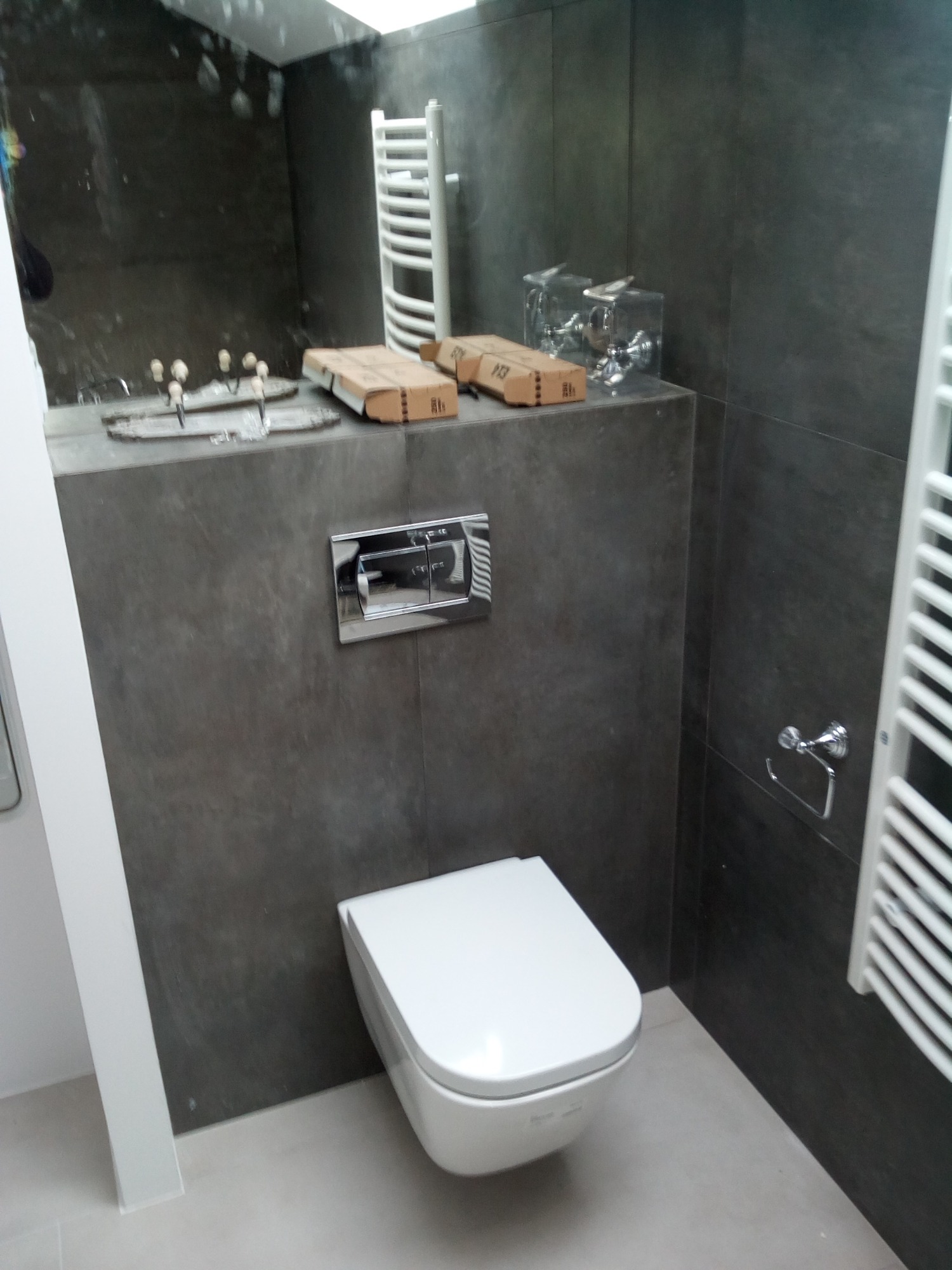 Nowoczesna toaleta podwieszana z chromowanym przyciskiem spłukującym na tle ciemnoszarych płytek, z widocznym grzejnikiem drabinkowym i akcesoriami łazienkowymi na blacie.