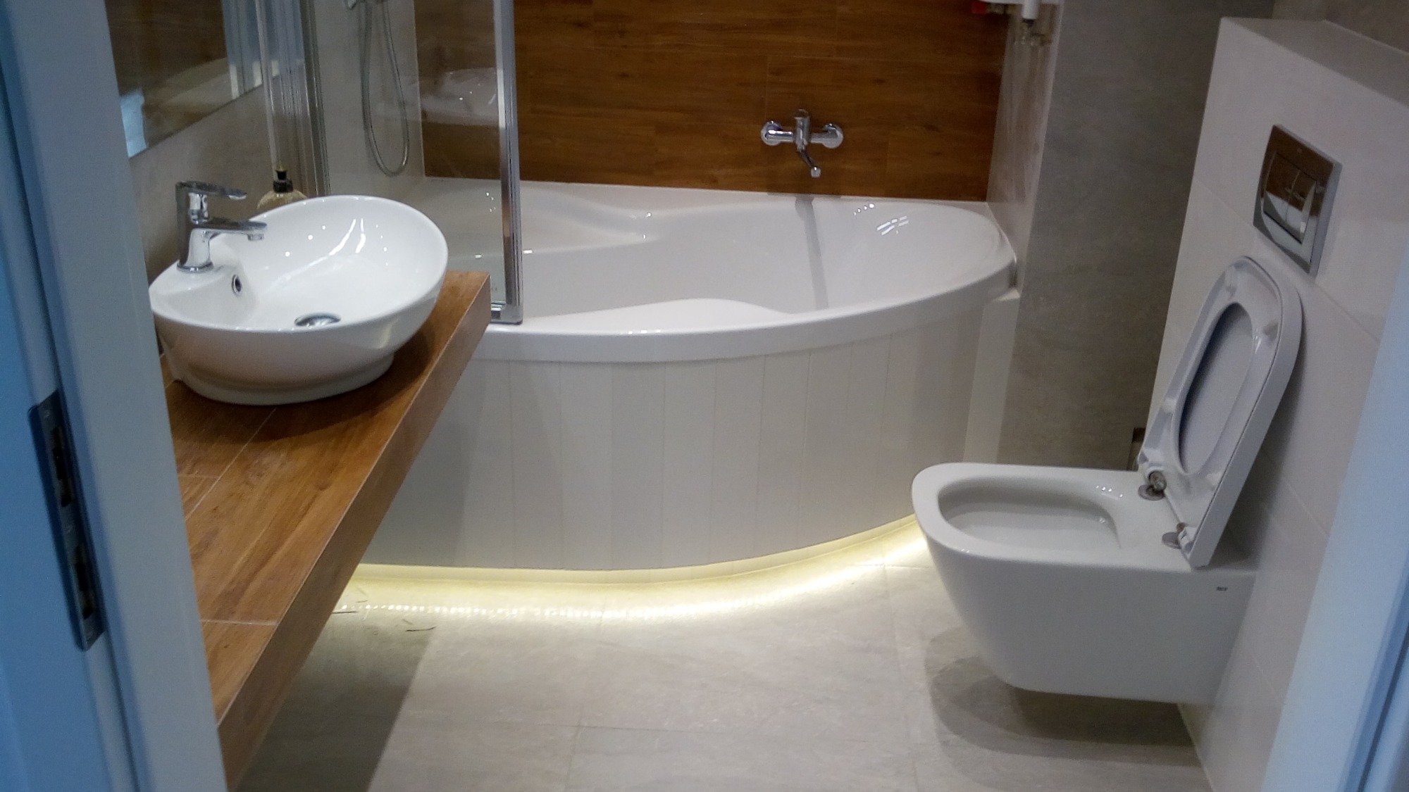 Nowoczesna łazienka z podświetloną wanną, umywalką na drewnianym blacie i podwieszaną toaletą.