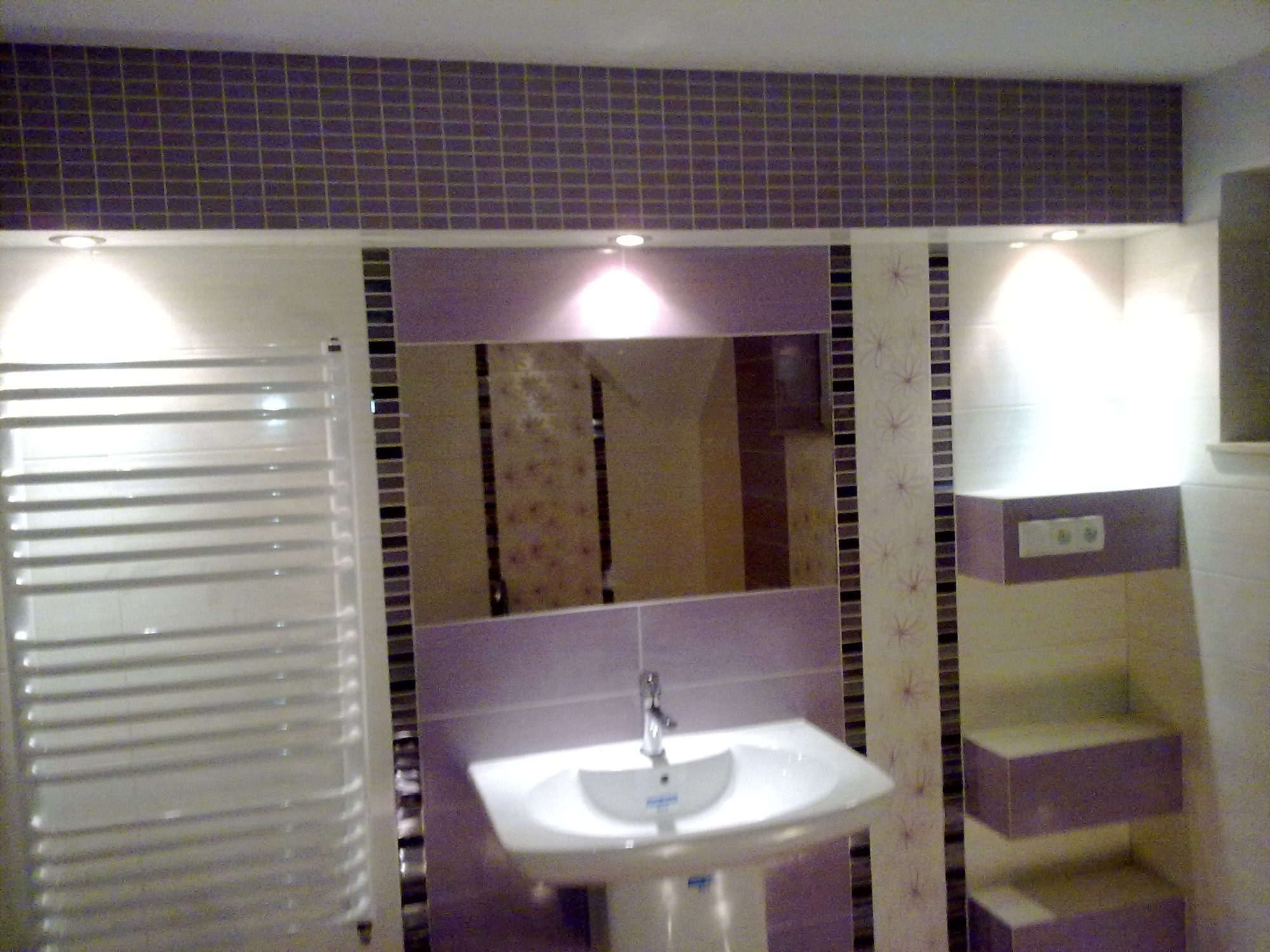 Wykończona łazienka z umywalką na postumencie, grzejnikiem drabinkowym i mozaiką w odcieniach fioletu i szarości podświetlona punktowymi lampami.