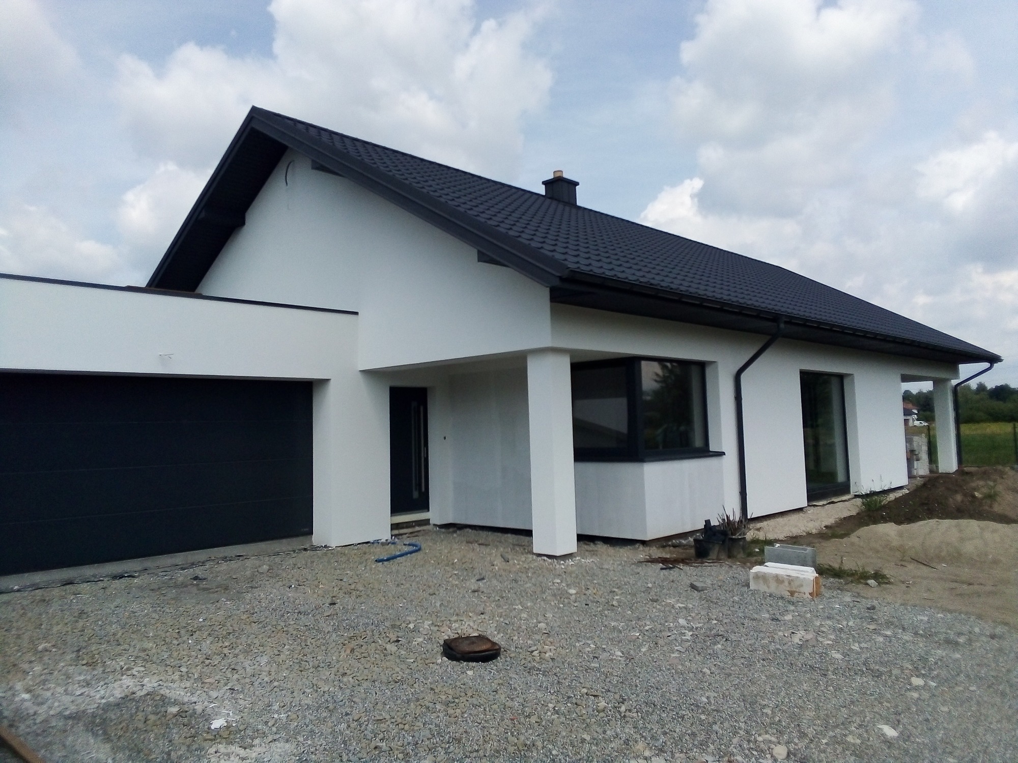 Nowoczesny dom jednorodzinny z ciemną dachówką i jasną elewacją w trakcie wykończenia terenu wokół budynku, widoczny podjazd wysypany szarym żwirem i materiały budowlane.