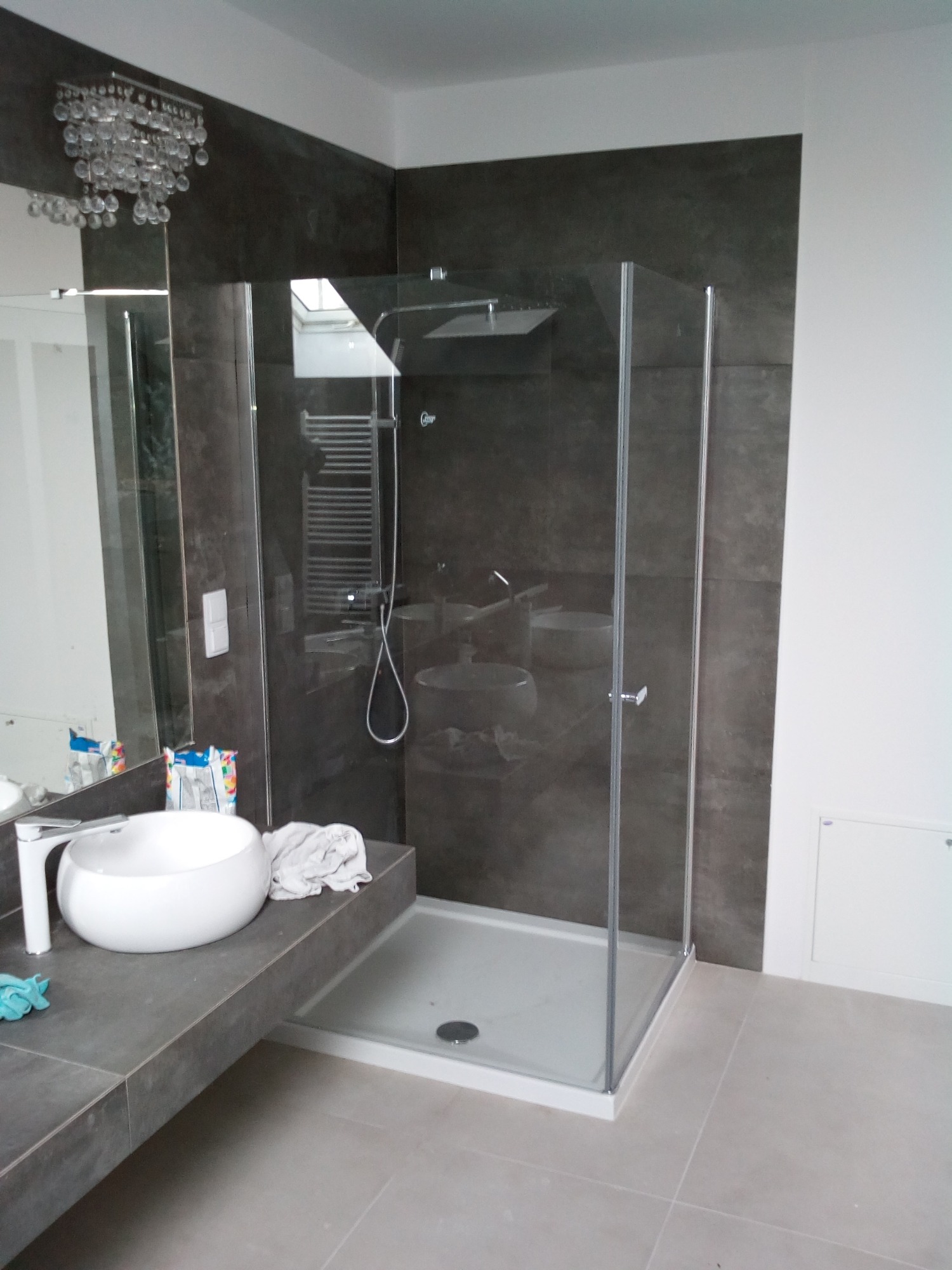 Nowoczesna łazienka z kabiną prysznicową typu walk-in, umywalką nablatową i grzejnikiem drabinkowym na tle ciemnoszarych, kwadratowych płytek. Nad umywalką widoczny ozdobny żyrandol.