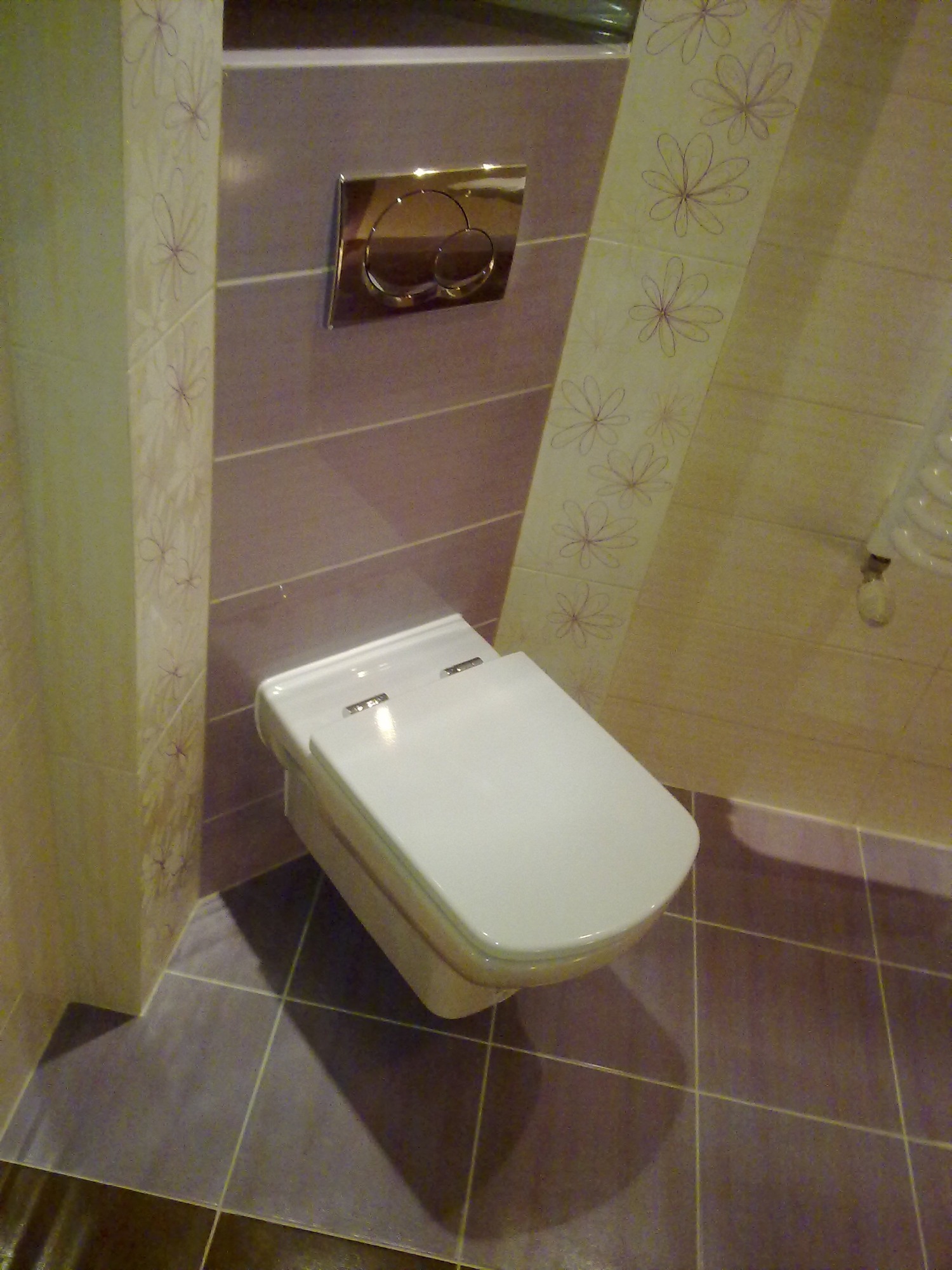 Nowoczesna toaleta podwieszana z chromowanym przyciskiem spłukującym, otoczona fioletowymi i kremowymi płytkami z motywem kwiatowym.