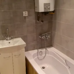 Nowoczesna łazienka z wanną, umywalką z szafką i wiszącym piecem gazowym Vaillant, wykończona szarymi płytkami ceramicznymi i drewnopodobną okładziną wanny.