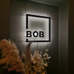Podświetlane logo 'BOB' w czarnej ramie na ścianie, z dekoracyjnym bukietem z suszonych traw i kwiatów w dolnej części kadru.
