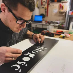 Mężczyzna w okularach precyzyjnie wykańcza czarny szyld z wyciętym, ozdobnym napisem 'Błogość' w warsztacie, używając małego narzędzia.