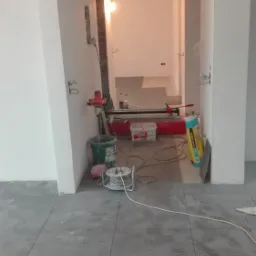 Korytarz w trakcie układania szarej terakoty, widoczne ślady butów, maszyna do cięcia płytek, wiadro z mieszadłem i puszka z klejem.