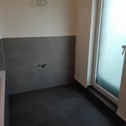 Częściowo wykończona łazienka z szarymi płytkami na ścianach i podłodze, widoczne przyłącza wodne i szyba prysznicowa z matowego szkła.