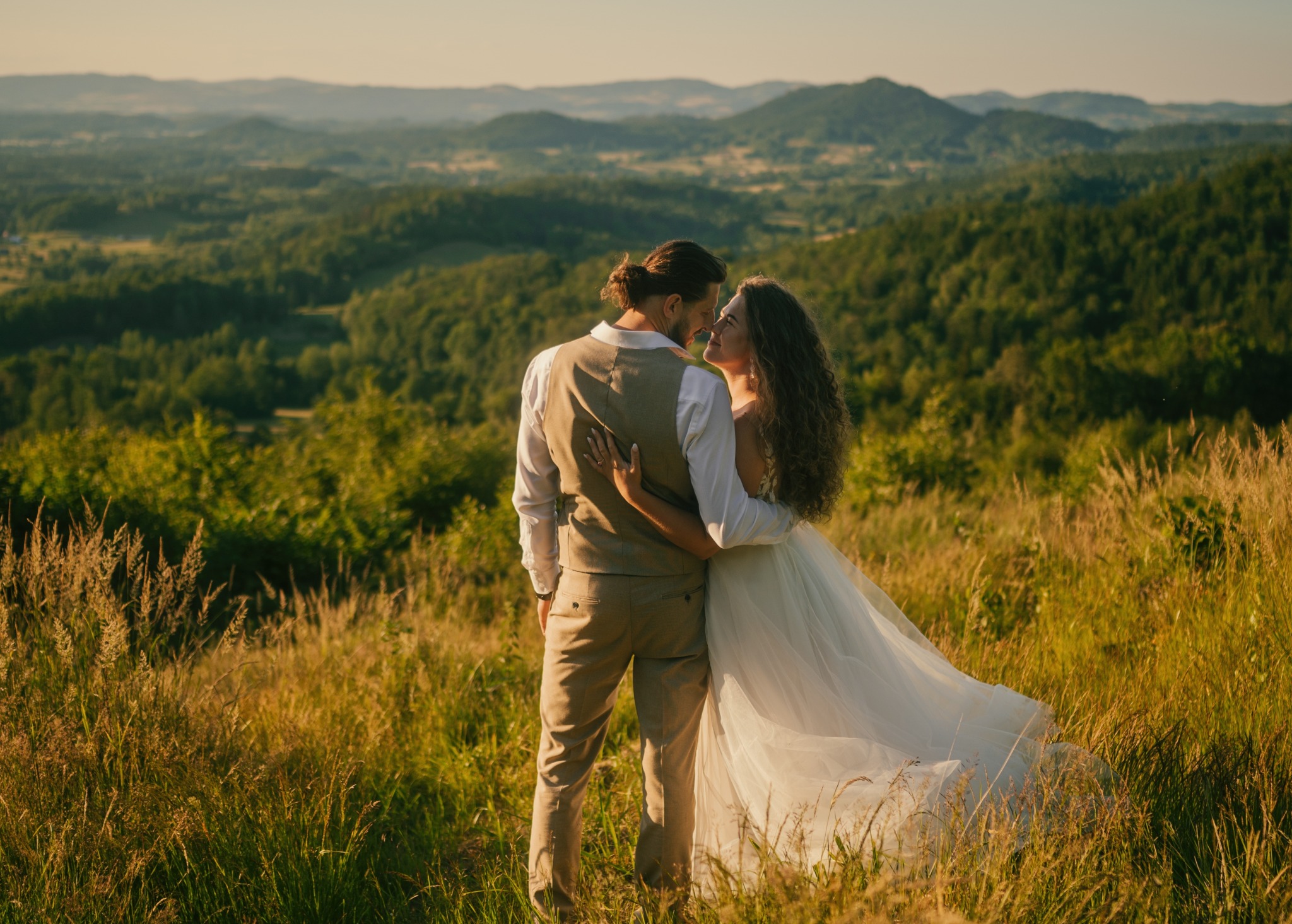 Para młoda w objęciach na tle rozległego krajobrazu górskiego o zachodzie słońca, panna młoda w zwiewnej sukni, pan młody w kamizelce i spodniach w kolorze beżowym, wysoka trawa w pierwszym planie.
