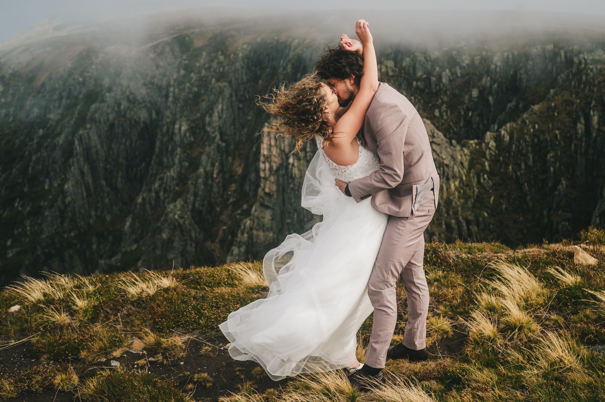 Para młoda w objęciach na tle górskiego krajobrazu; panna młoda w białej sukni z koronkową górą, pan młody w jasnobrązowym garniturze, w tle skaliste szczyty spowite mgłą.