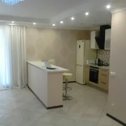 Jasna kuchnia z wyspą i szarymi, kwadratowymi płytkami podłogowymi, widok na wyspę z laptopem, hoker i fragment zabudowy kuchennej z AGD.