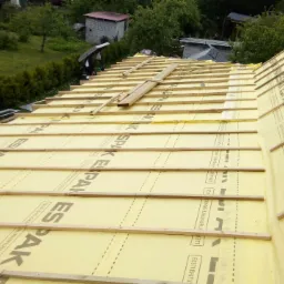 Montaż izolacji termicznej ESPAK na dachu z widocznymi łatami drewnianymi, w tle zieleń drzew i budynki gospodarcze.