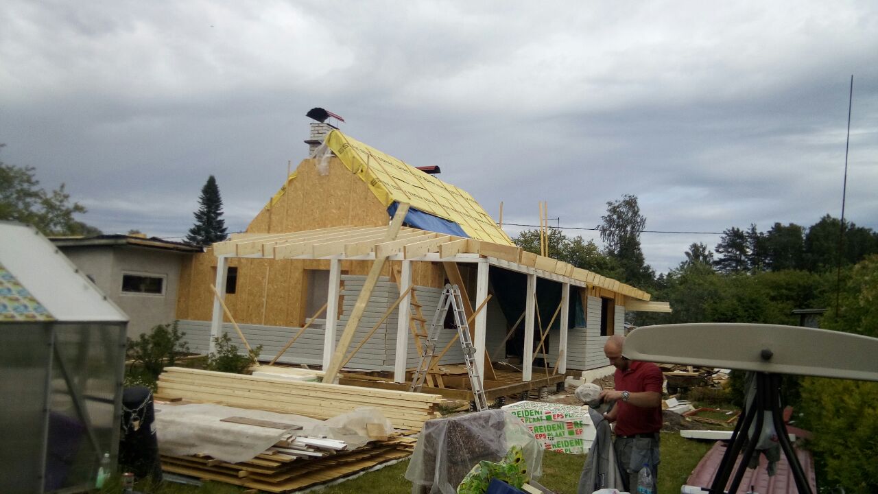 Budowa domu w trakcie: szkielet drewniany, płyty OSB, żółta membrana dachowa, widoczny mężczyzna w czerwonej koszulce pracujący przy elewacji, w tle zieleń ogrodu i pochmurne niebo.