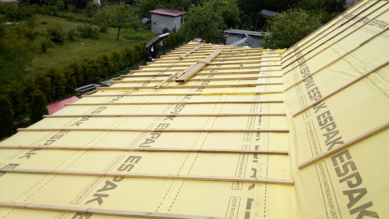 Montaż izolacji termicznej ESPAK na dachu z widocznymi łatami drewnianymi, w tle zieleń drzew i budynki gospodarcze.