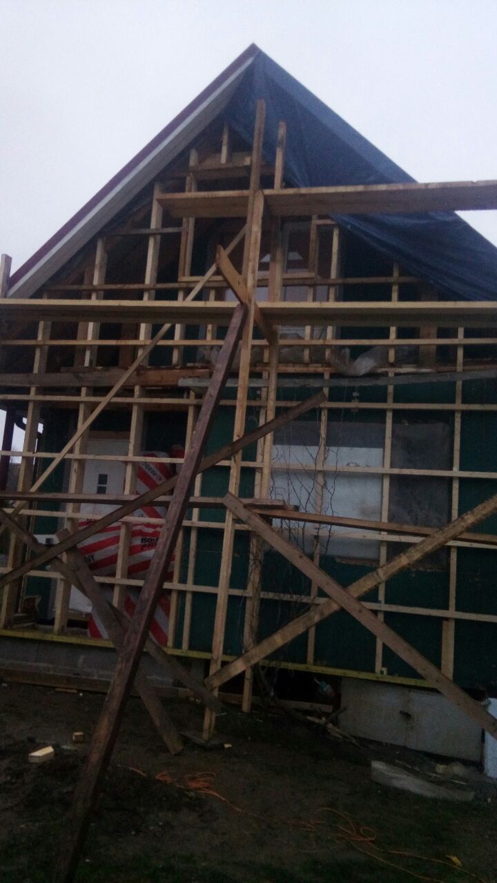 Konstrukcja szkieletowa domu w trakcie budowy, widoczne drewniane belki i częściowo ocieplone ściany, na dachu plandeka ochronna.