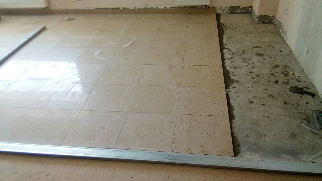 Układanie jasnych, kwadratowych płytek podłogowych w pomieszczeniu, widoczna listwa przypodłogowa i fragment surowego podłoża przed wyłożeniem.