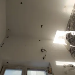 Poddasze w trakcie prac wykończeniowych: widoczne otwory na oświetlenie w ścianie, zabezpieczone okno folią malarską, fragment instalacji elektrycznej i plama światła słonecznego.